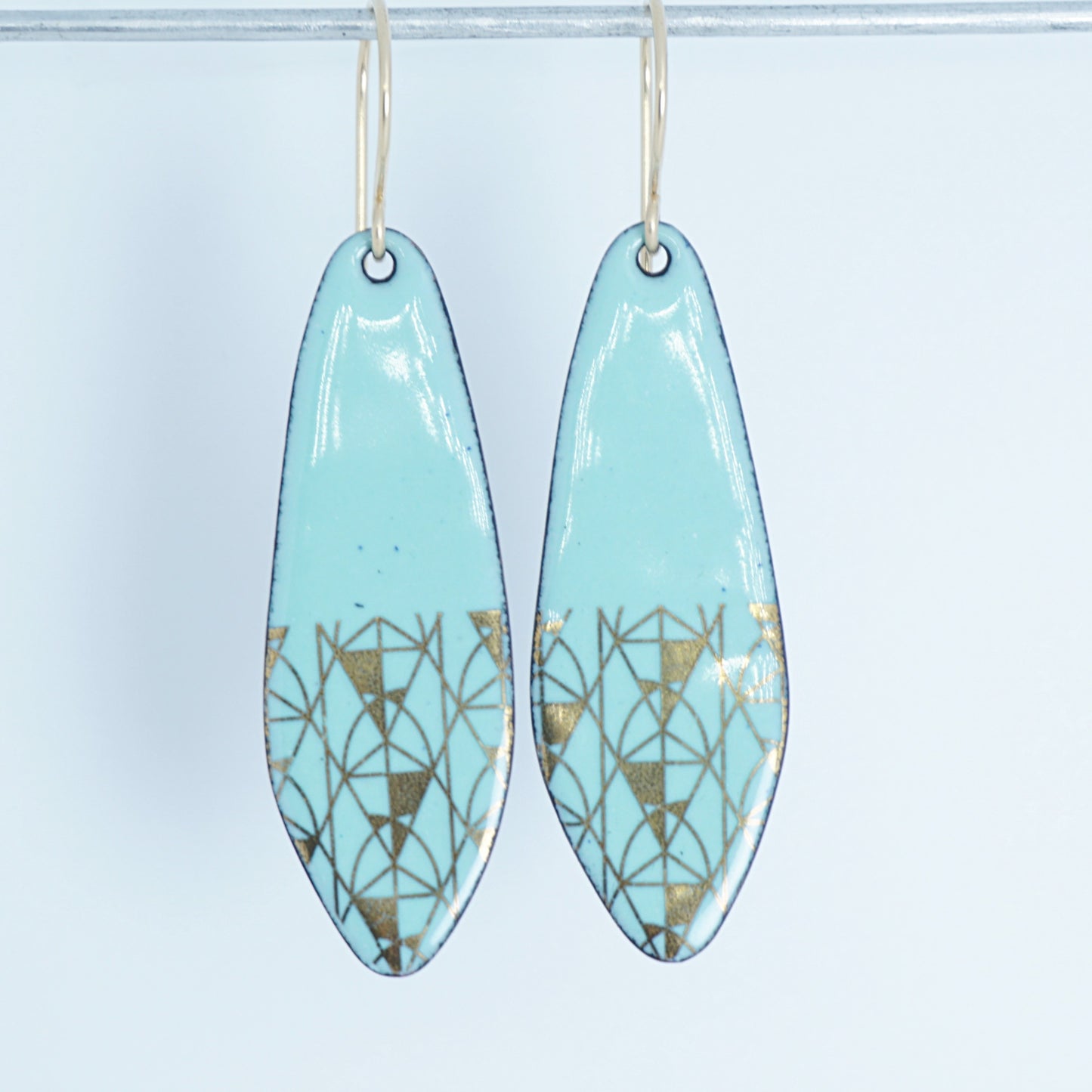 A pair of seafoam green teardrop shaped earrings with gold geometric accents, suspended from thin gold hooks on a white background