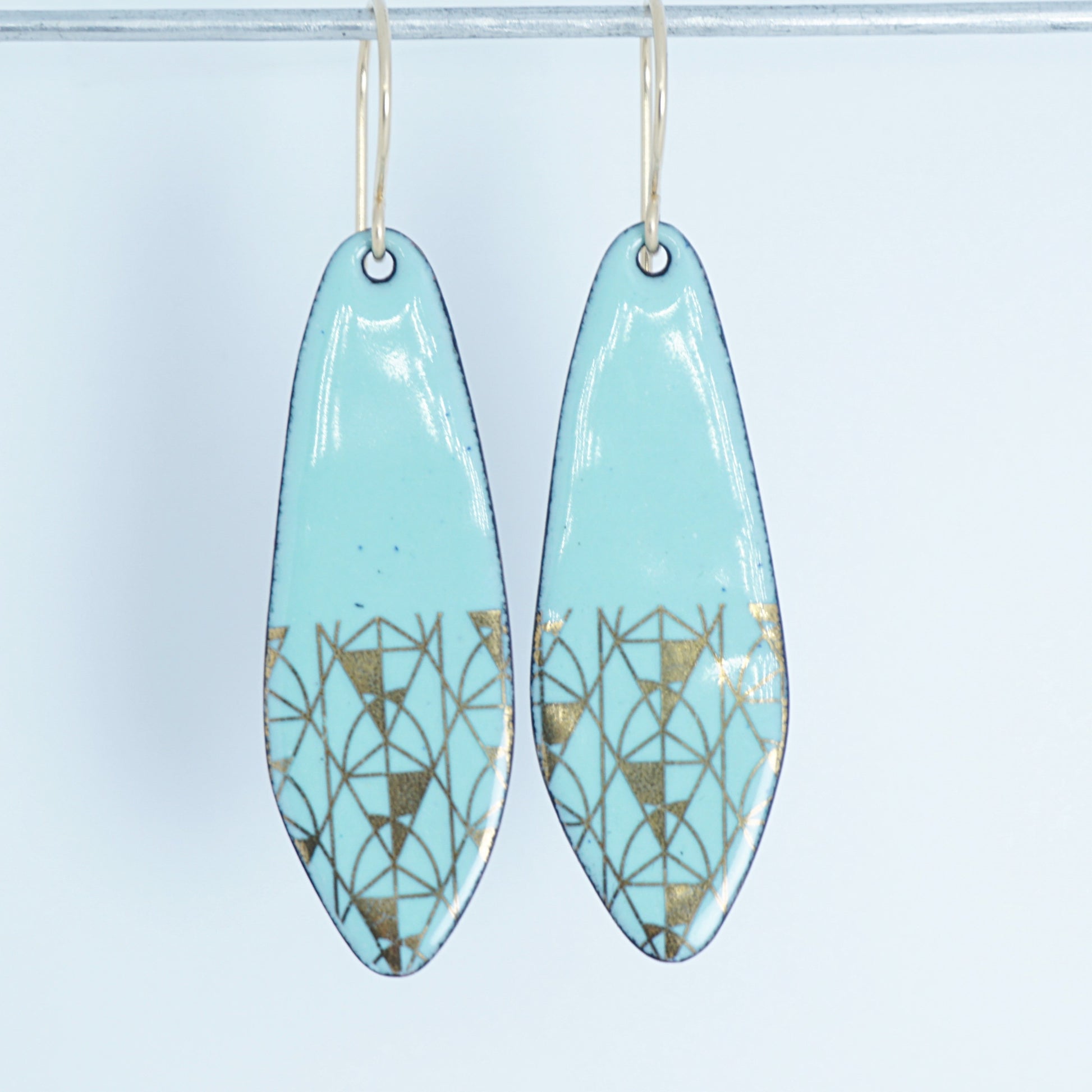 A pair of seafoam green teardrop shaped earrings with gold geometric accents, suspended from thin gold hooks on a white background