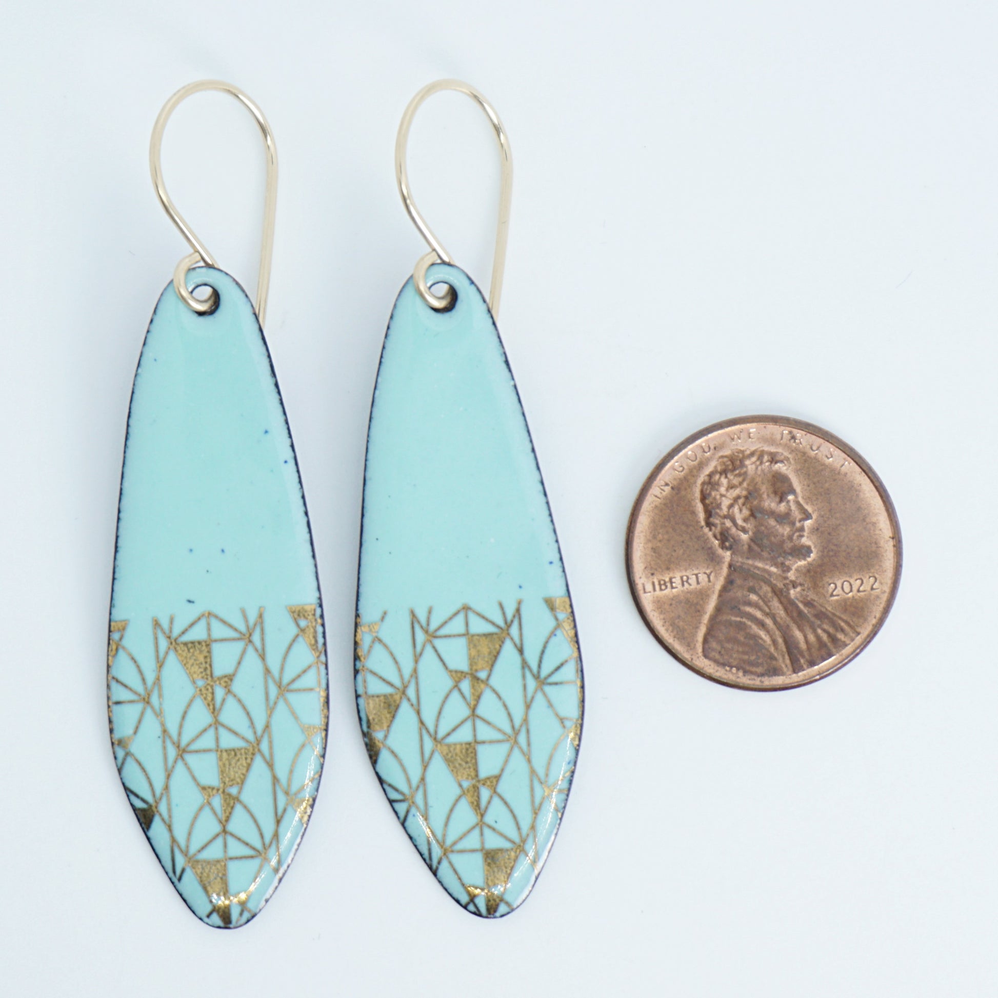 A pair of seafoam green teardrop earrings with gold geometric line patterns near the bottom, shown next to a penny for scale, against a white background