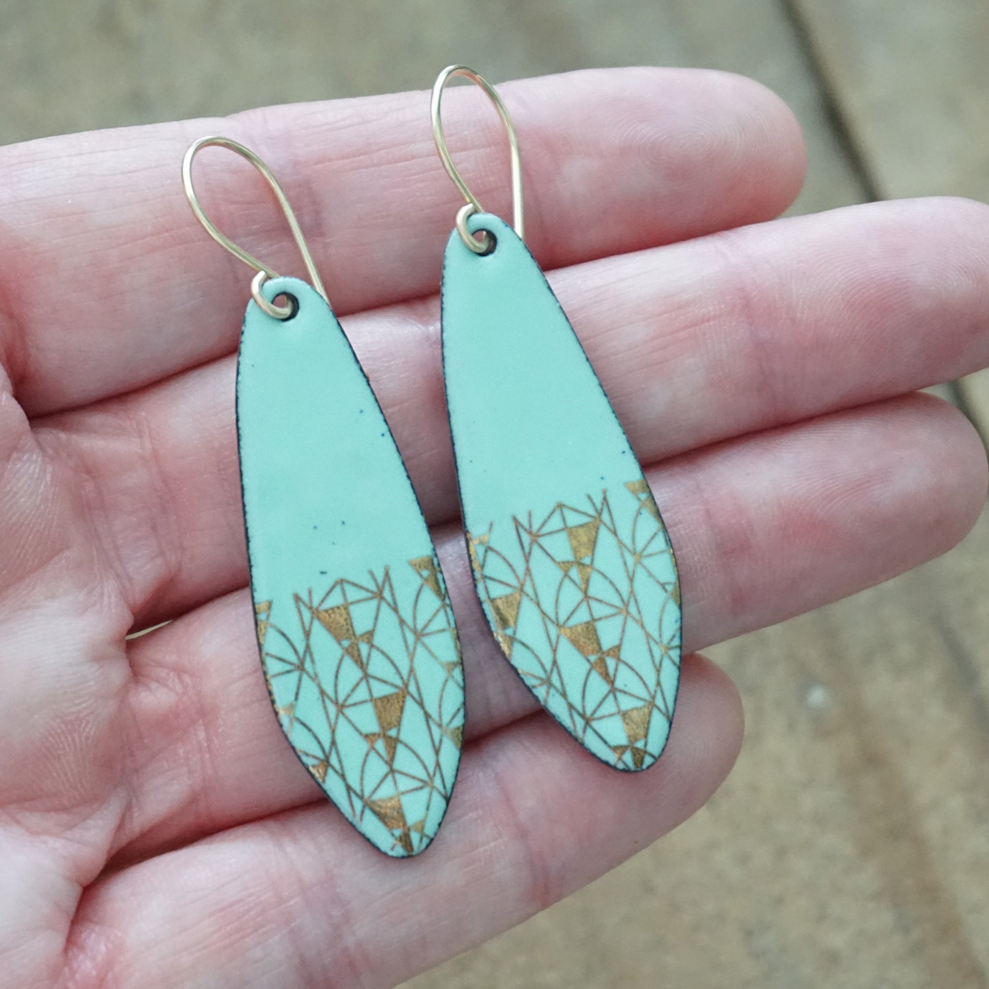 A pair of dangling earrings held in a person's hand. The earrings have a mint green base with a gold geometric triangle pattern near the bottom and gold fishhook ear wires.