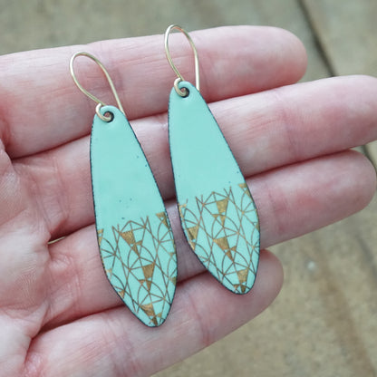 A pair of dangling earrings held in a person's hand. The earrings have a mint green base with a gold geometric triangle pattern near the bottom and gold fishhook ear wires.