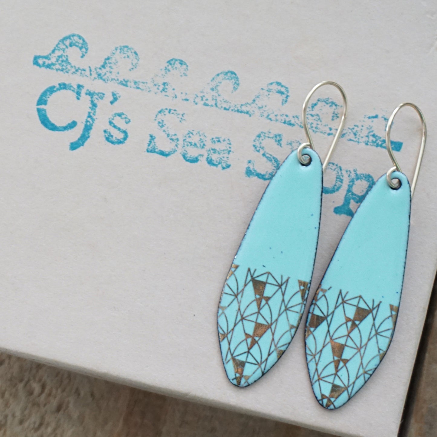 A pair of seafoam green teardrop shaped earrings with gold geometric accents, suspended from thin gold hooks on a gift box