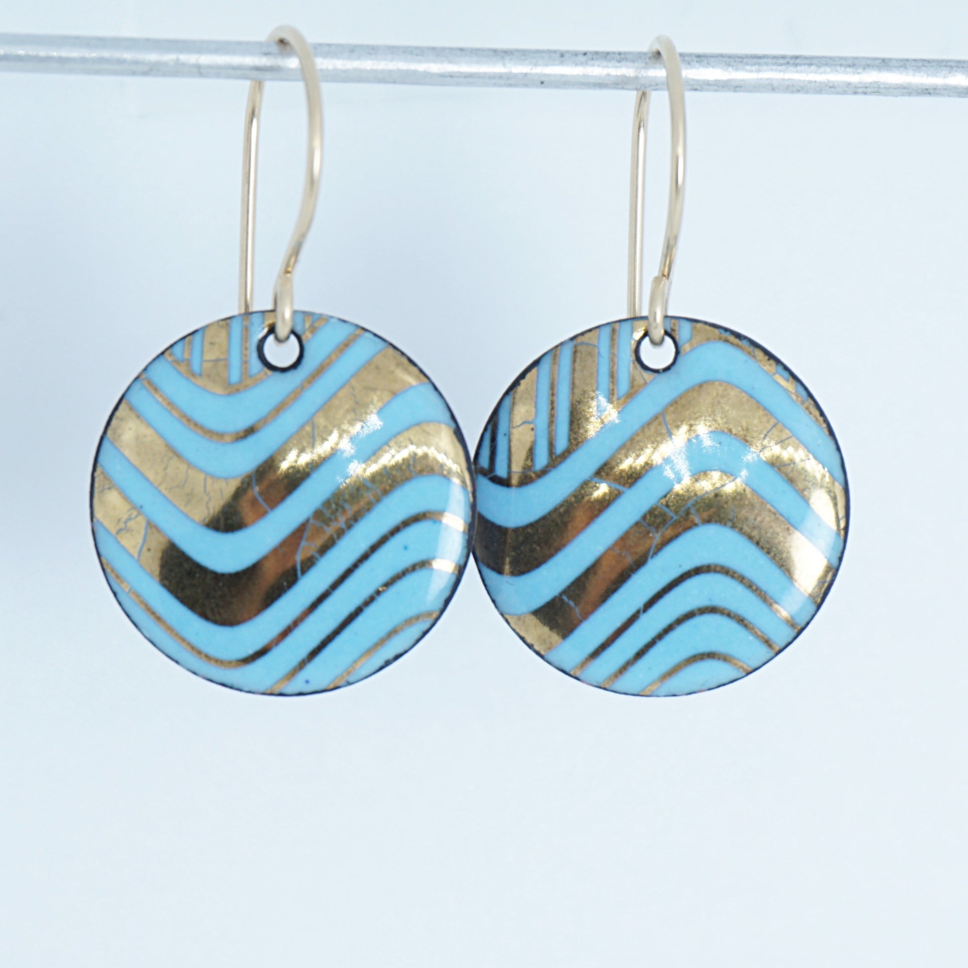 A pair of round drop earrings with light blue and gold wavy patterned enamel and gold-tone fishhook wires, shown on a white background