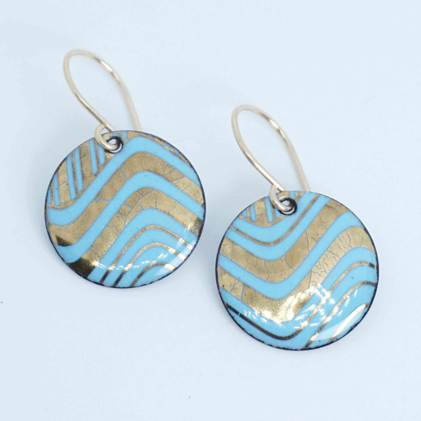 A pair of round drop earrings with light blue and gold wavy patterned enamel and gold-tone fishhook wires, shown on a white background