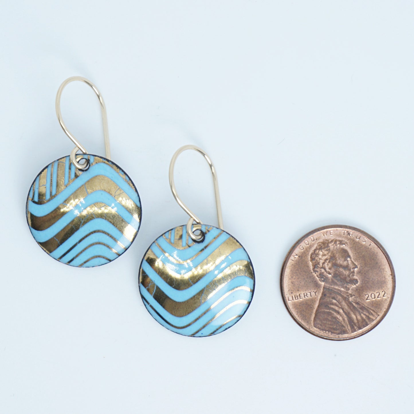 Two round drop earrings with a light blue and gold wavy pattern, shown next to a penny for scale on a white background.