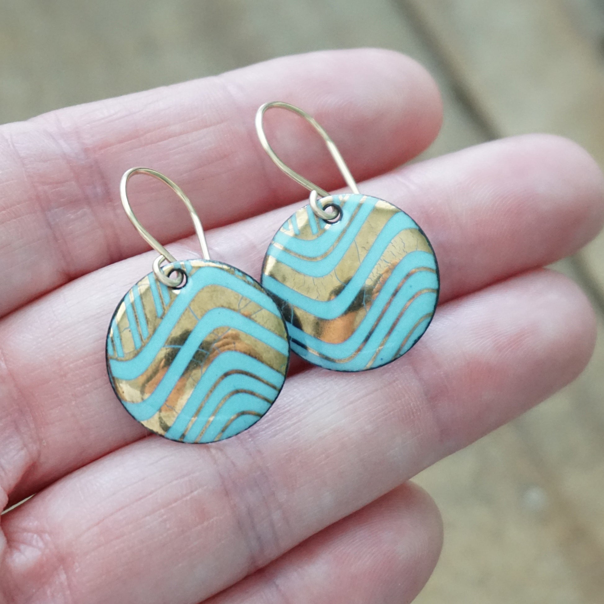 Hand holding a pair of round drop earrings with light blue and gold wavy patterned enamel and gold-tone fishhook wires, shown on a wooden background