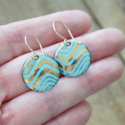 Hand holding a pair of round drop earrings with light blue and gold wavy patterned enamel and gold-tone fishhook wires, shown on a wooden background