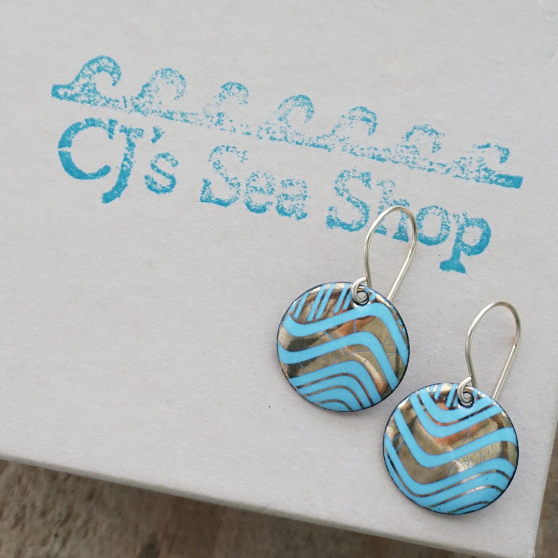 A pair of round drop earrings with light blue and gold wavy patterned enamel and gold-tone fishhook wires, shown on a gift box