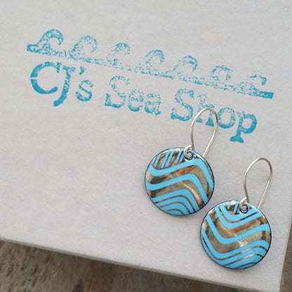 A pair of round drop earrings with light blue and gold wavy patterned enamel and gold-tone fishhook wires, shown on a gift box