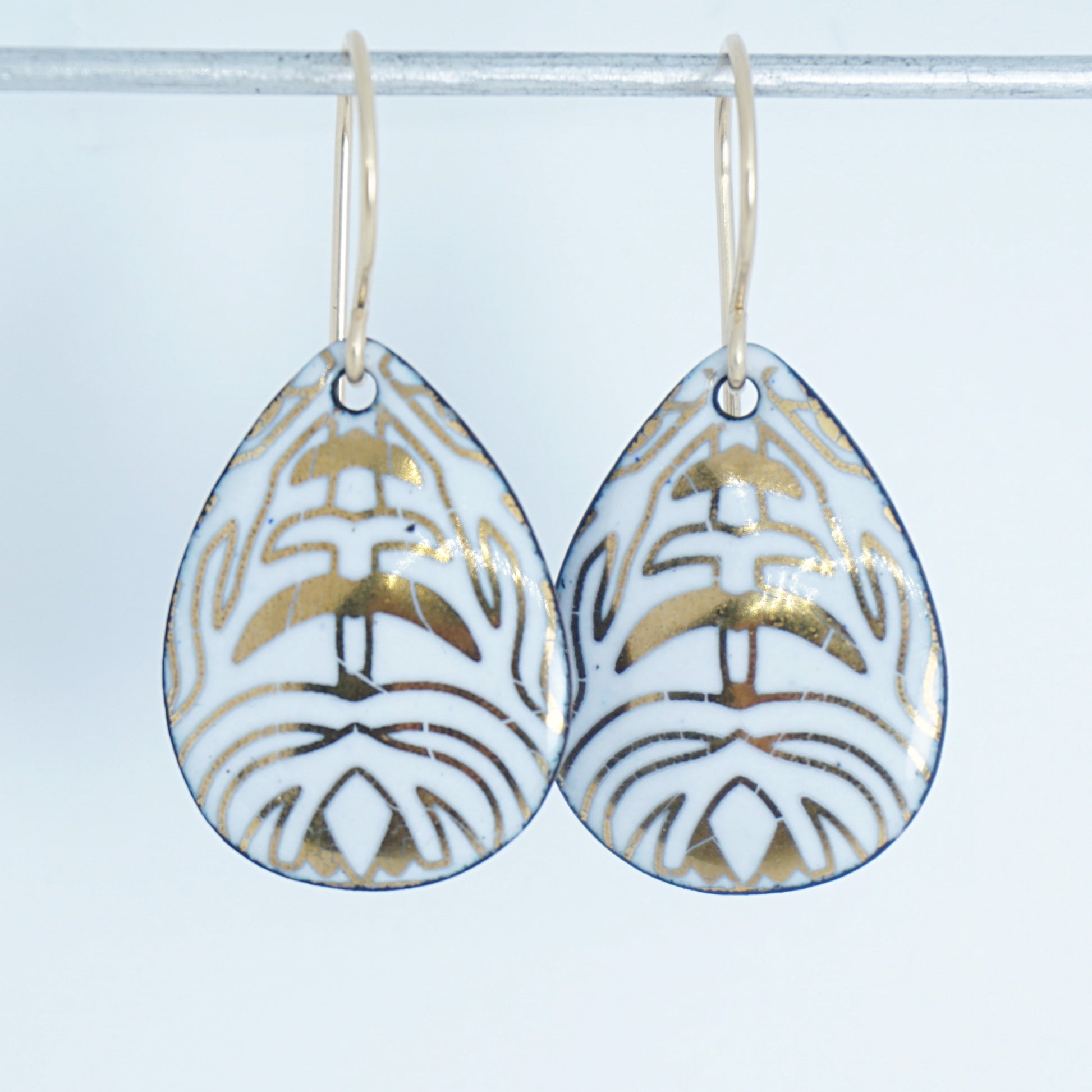 A pair of teardrop-shaped drop earrings with a white enamel background, gold leaf-like patterns, hanging from gold fishhook wires.