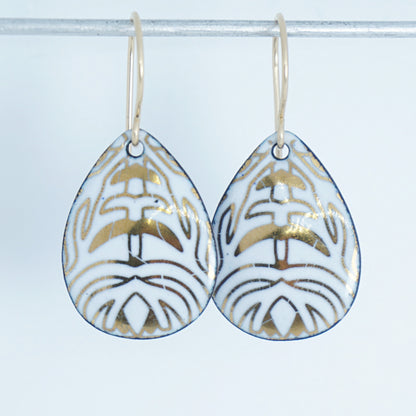 A pair of teardrop-shaped drop earrings with a white enamel background, gold leaf-like patterns, hanging from gold fishhook wires.
