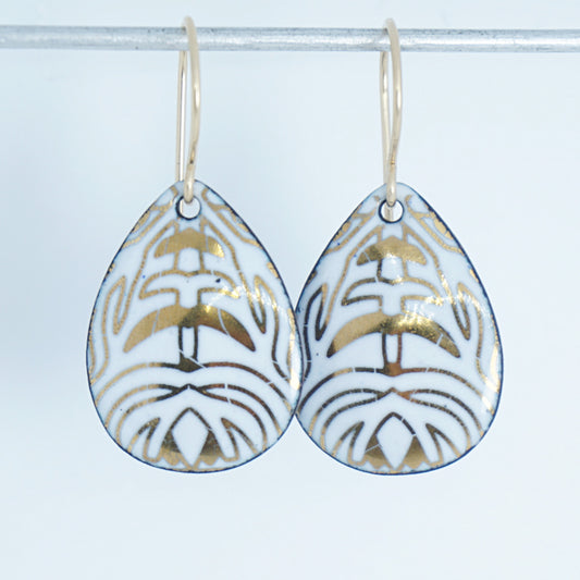 A pair of teardrop-shaped drop earrings with a white enamel background, gold leaf-like patterns, hanging from gold fishhook wires.