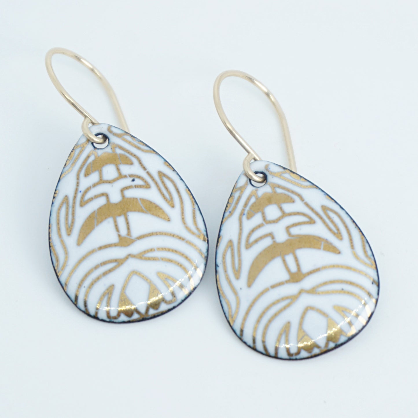 A pair of teardrop-shaped drop earrings with a white enamel background, gold leaf-like patterns, hanging from gold fishhook wires.