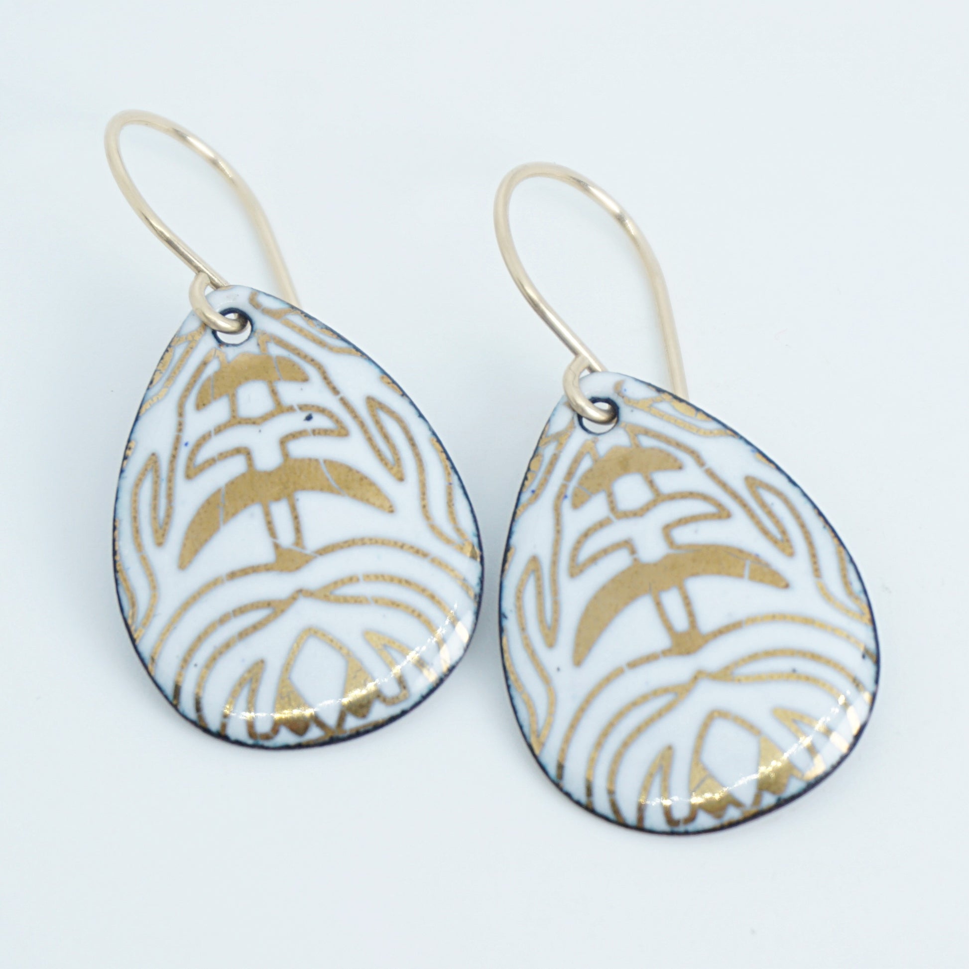 A pair of teardrop-shaped drop earrings with a white enamel background, gold leaf-like patterns, hanging from gold fishhook wires.