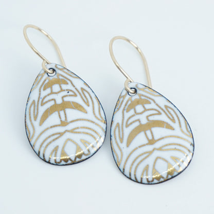 A pair of teardrop-shaped drop earrings with a white enamel background, gold leaf-like patterns, hanging from gold fishhook wires.