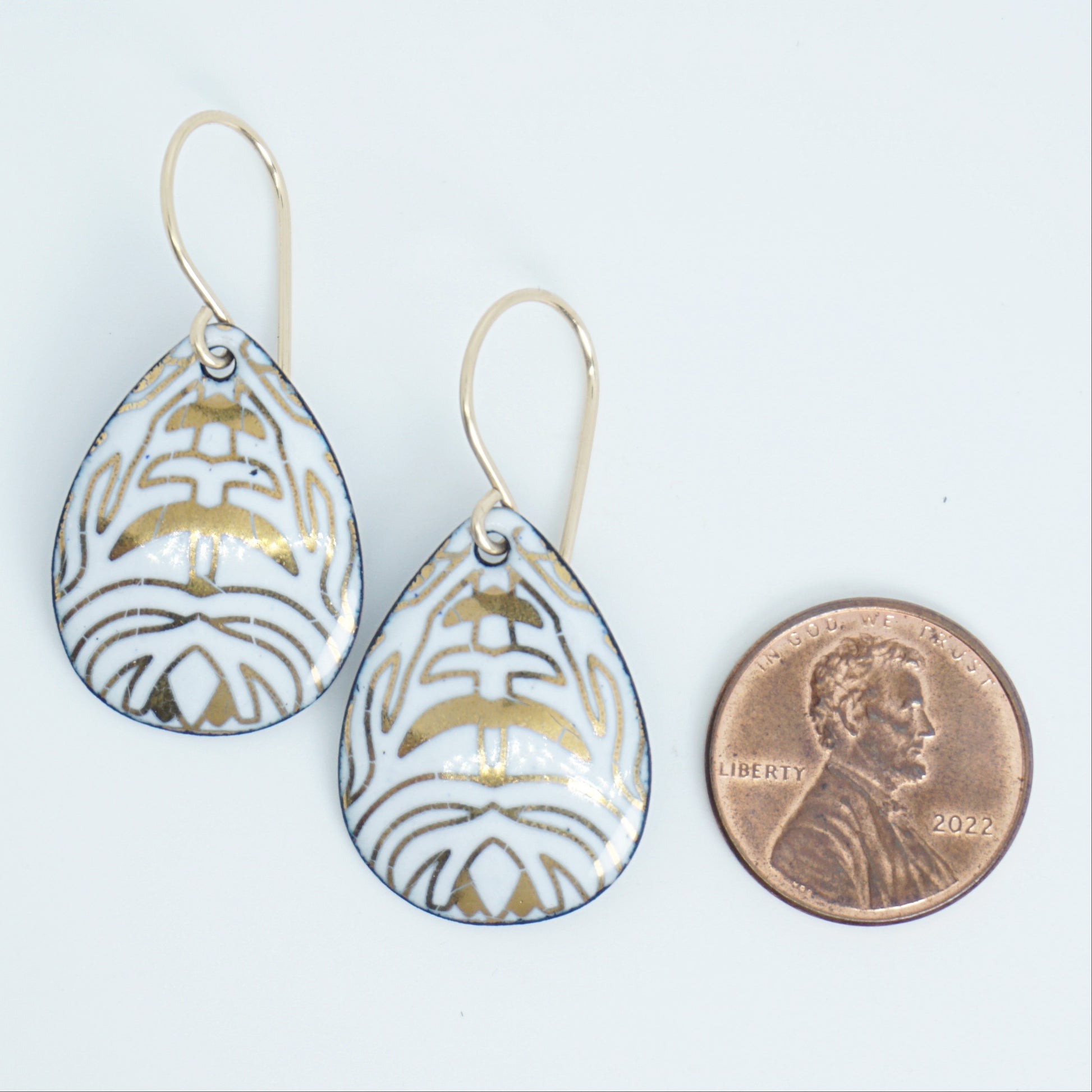 A pair of teardrop-shaped drop earrings with a white enamel background, gold leaf-like patterns, hanging from gold fishhook wires, next to a penny for scale, against a white background