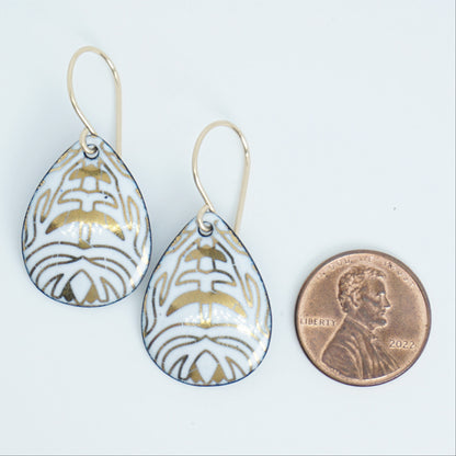 A pair of teardrop-shaped drop earrings with a white enamel background, gold leaf-like patterns, hanging from gold fishhook wires, next to a penny for scale, against a white background