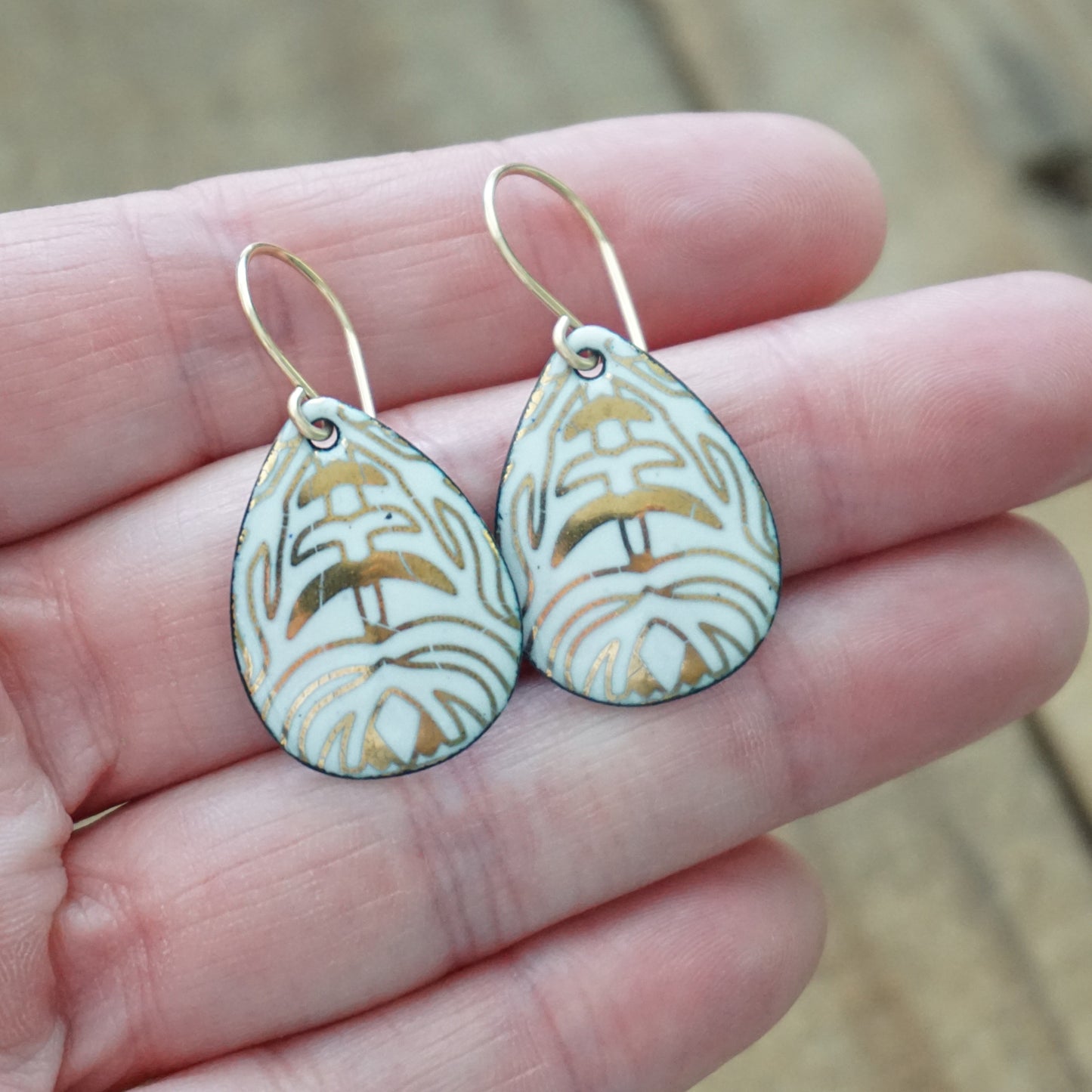 Hand holding A pair of teardrop-shaped drop earrings with a white enamel background, gold leaf-like patterns, hanging from gold fishhook wires, against a wooden background