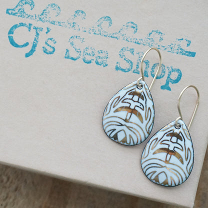 A pair of teardrop-shaped drop earrings with a white enamel background, gold leaf-like patterns, hanging from gold fishhook wires, on a gift box