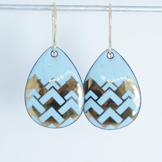 A pair of teardrop-shaped dangle earrings with a light blue enamel background, featuring a metallic gold  chevron pattern, hanging from thin gold hooks.
