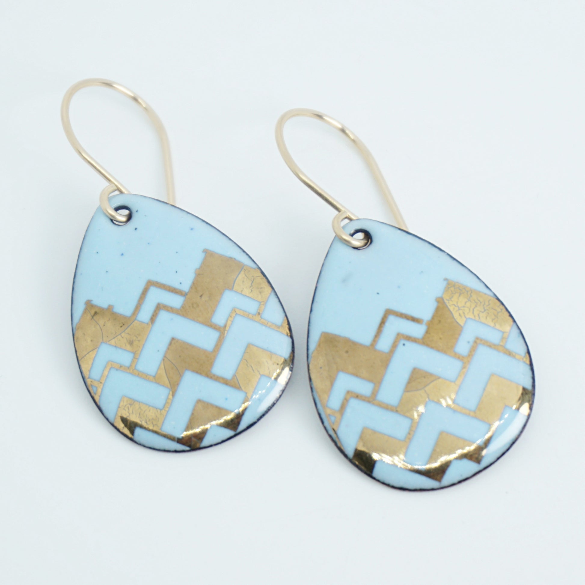 A pair of teardrop-shaped dangle earrings with a light blue enamel background, featuring a metallic gold  chevron pattern, hanging from thin gold hooks.