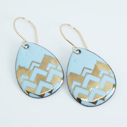 A pair of teardrop-shaped dangle earrings with a light blue enamel background, featuring a metallic gold  chevron pattern, hanging from thin gold hooks.
