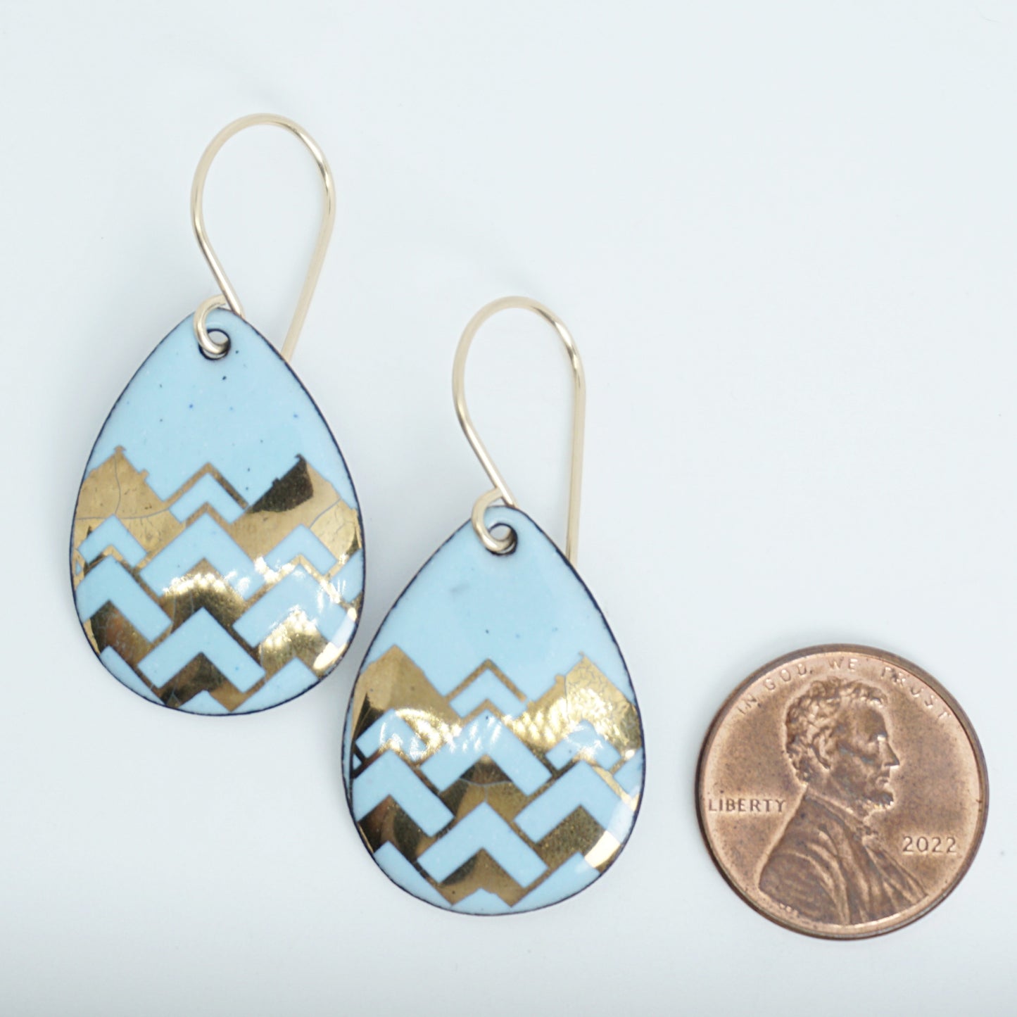 A pair of teardrop-shaped dangle earrings with a light blue enamel background, featuring a metallic gold  chevron pattern, hanging from thin gold hooks, next to a penny for scale, against a white background