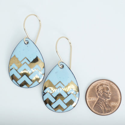 A pair of teardrop-shaped dangle earrings with a light blue enamel background, featuring a metallic gold  chevron pattern, hanging from thin gold hooks, next to a penny for scale, against a white background