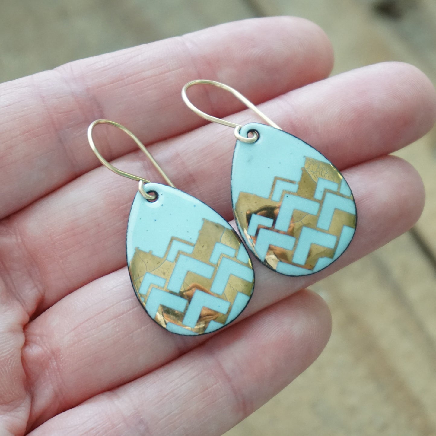 Hand holding A pair of teardrop-shaped dangle earrings with a light blue enamel background, featuring a metallic gold  chevron pattern, hanging from thin gold hooks.