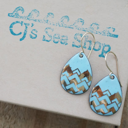 A pair of teardrop-shaped dangle earrings with a light blue enamel background, featuring a metallic gold  chevron pattern, hanging from thin gold hooks, displayed on a gift box