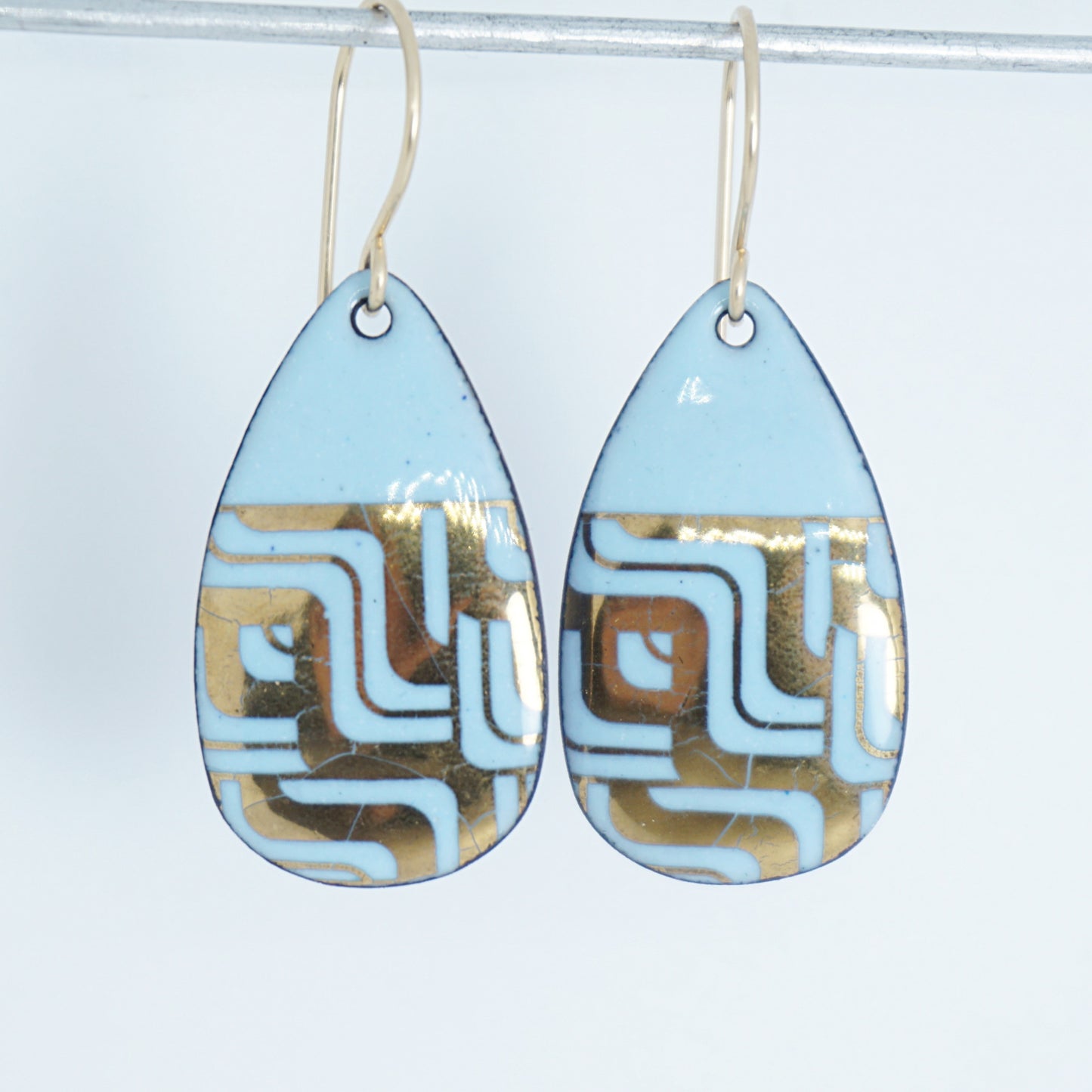 A pair of teardrop-shaped dangle earrings with a light blue enamel background, featuring a metallic geometric pattern, hanging from thin gold hooks.