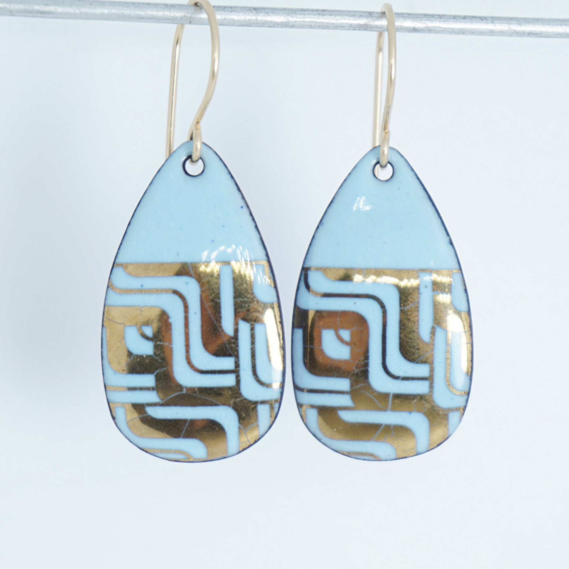 A pair of teardrop-shaped dangle earrings with a light blue enamel background, featuring a metallic geometric pattern, hanging from thin gold hooks.
