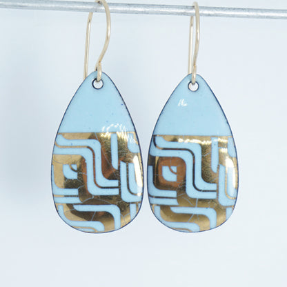 A pair of teardrop-shaped dangle earrings with a light blue enamel background, featuring a metallic geometric pattern, hanging from thin gold hooks.