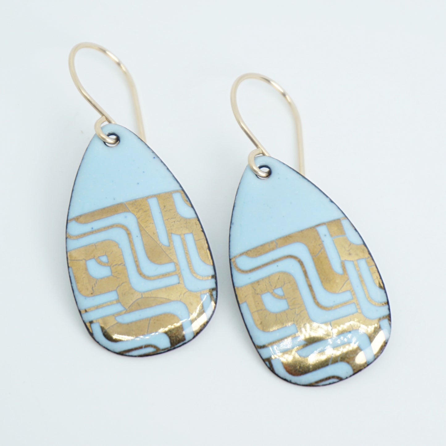 A pair of teardrop-shaped dangle earrings with a light blue enamel background, featuring a metallic geometric pattern, hanging from thin gold hooks.