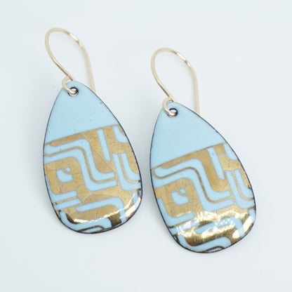 A pair of teardrop-shaped dangle earrings with a light blue enamel background, featuring a metallic geometric pattern, hanging from thin gold hooks.