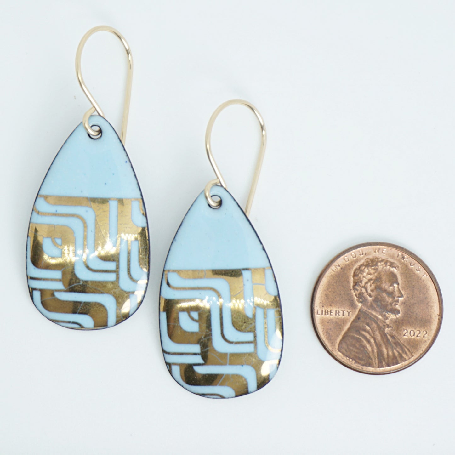 A pair of teardrop-shaped dangle earrings with a light blue enamel background, featuring a metallic geometric pattern, hanging from thin gold hooks, next to a penny for scale, against a white background
