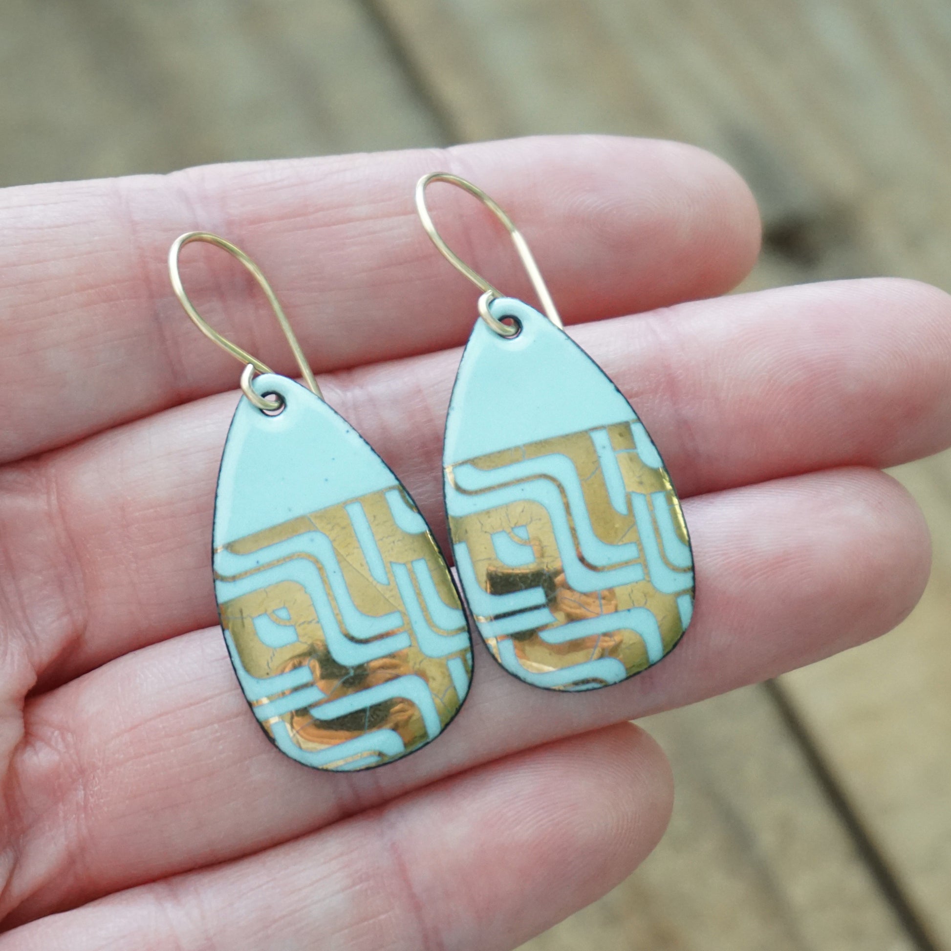 Hand holding A pair of teardrop-shaped dangle earrings with a light blue enamel background, featuring a metallic geometric pattern, hanging from thin gold hooks, against a wooden background