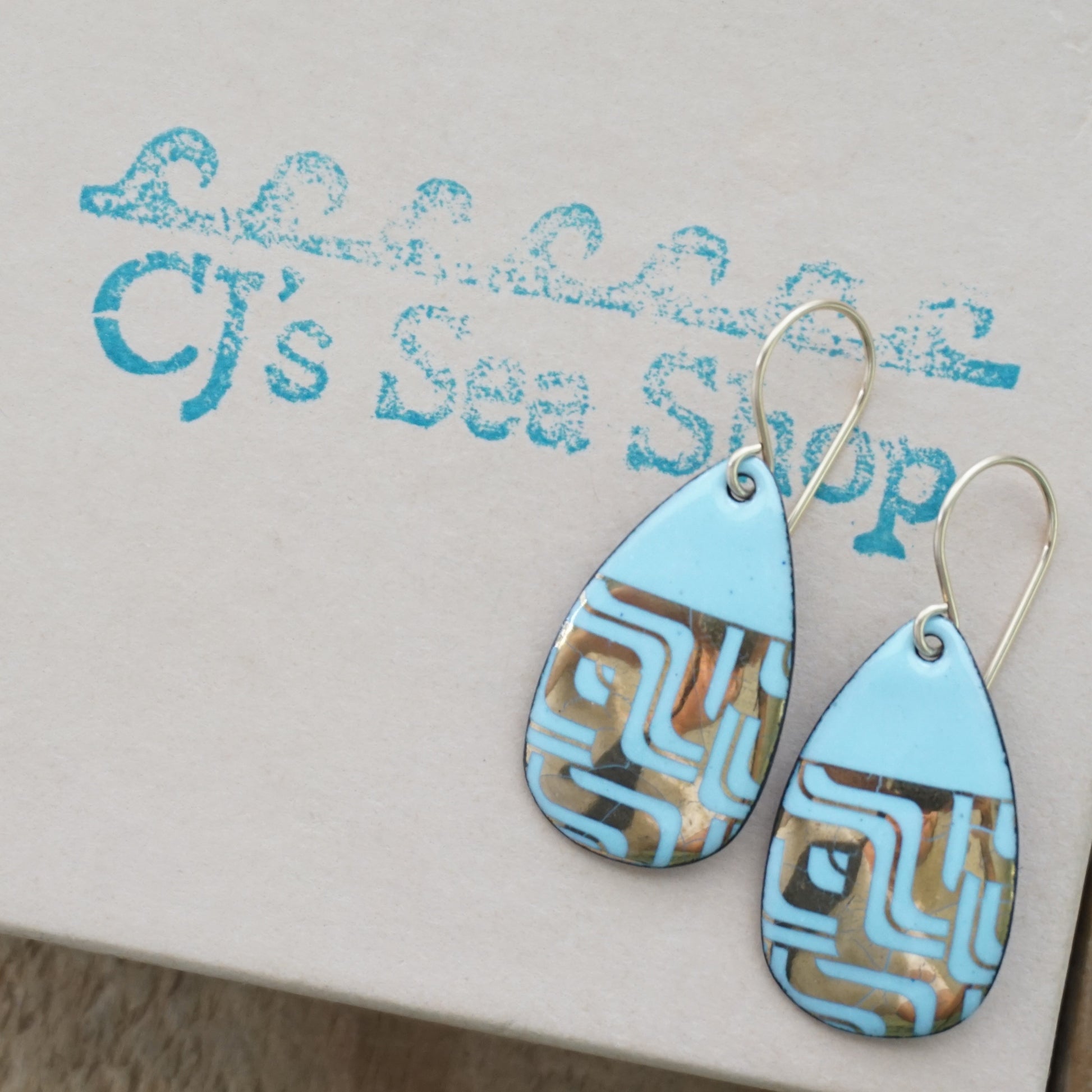 A pair of teardrop-shaped dangle earrings with a light blue enamel background, featuring a metallic geometric pattern, hanging from thin gold hooks, displayed on a gift box