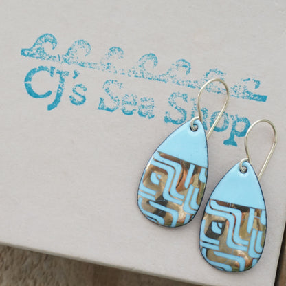 A pair of teardrop-shaped dangle earrings with a light blue enamel background, featuring a metallic geometric pattern, hanging from thin gold hooks, displayed on a gift box