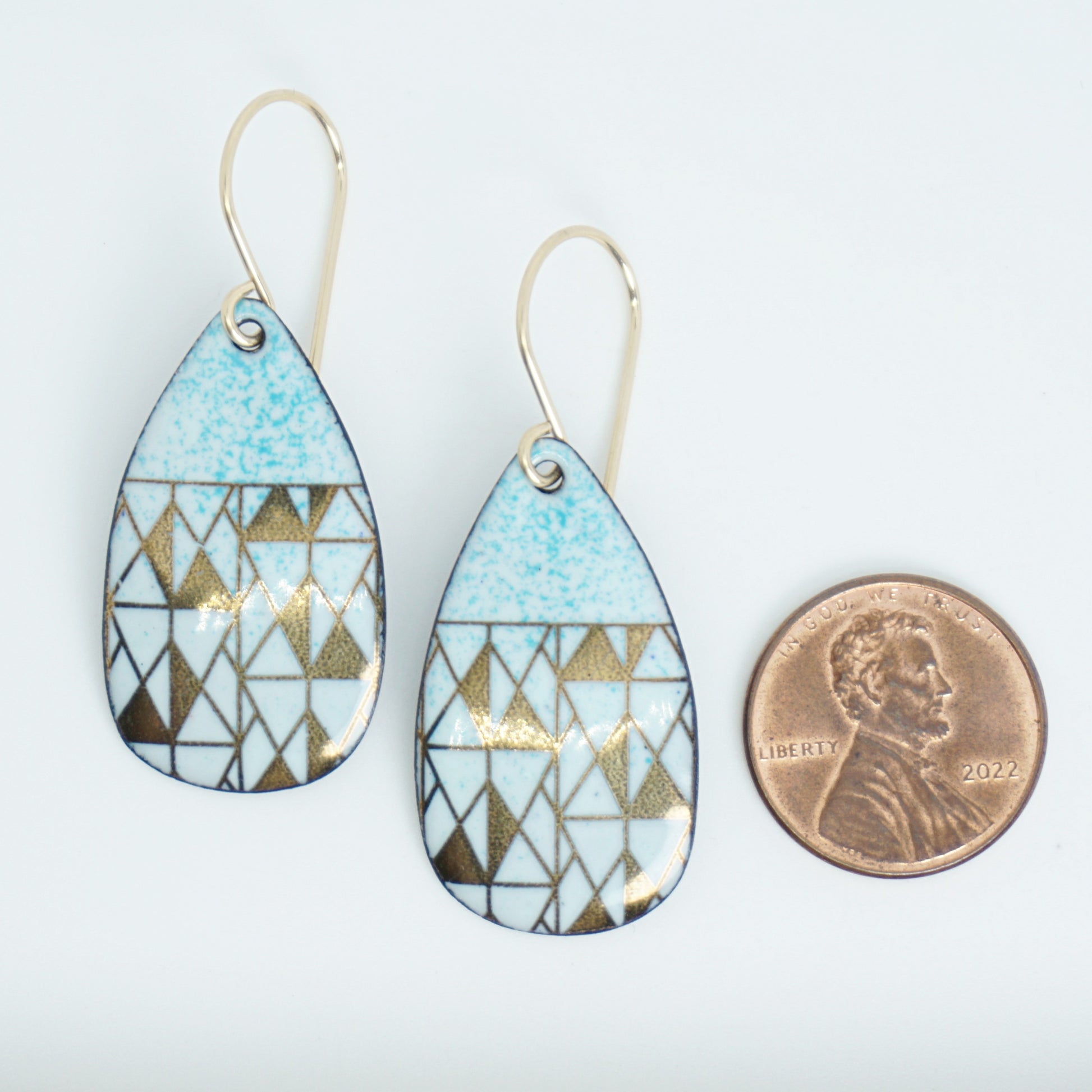 A pair of teardrop-shaped dangle earrings with seafoam green enamel and gold geometric pattern, next to a penny for scale, against a white background