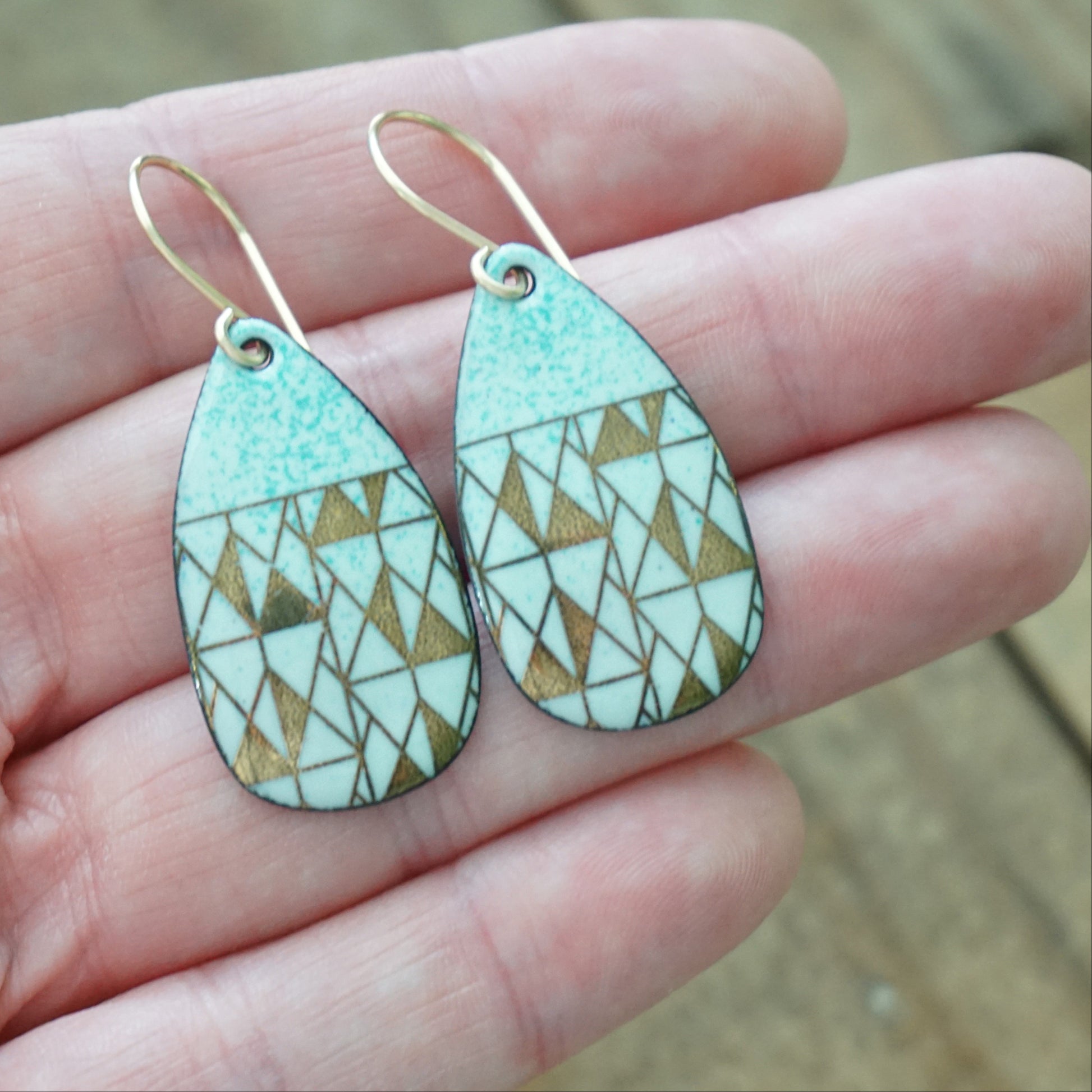 hand holding A pair of teardrop-shaped dangle earrings with seafoam green enamel and gold geometric pattern, against a wooden background