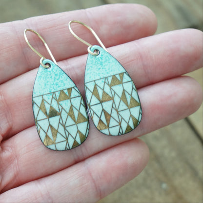 hand holding A pair of teardrop-shaped dangle earrings with seafoam green enamel and gold geometric pattern, against a wooden background