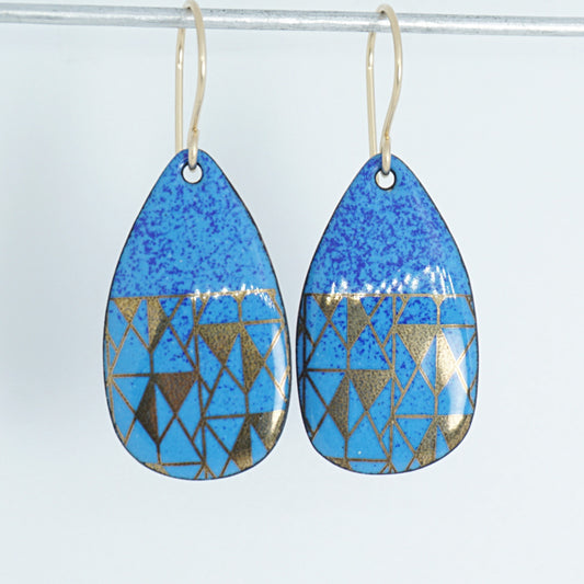 A pair of teardrop-shaped drop earrings with a blue speckled top half and a geometric gold triangle pattern on a blue lower half, hanging from thin gold hooks against a white background