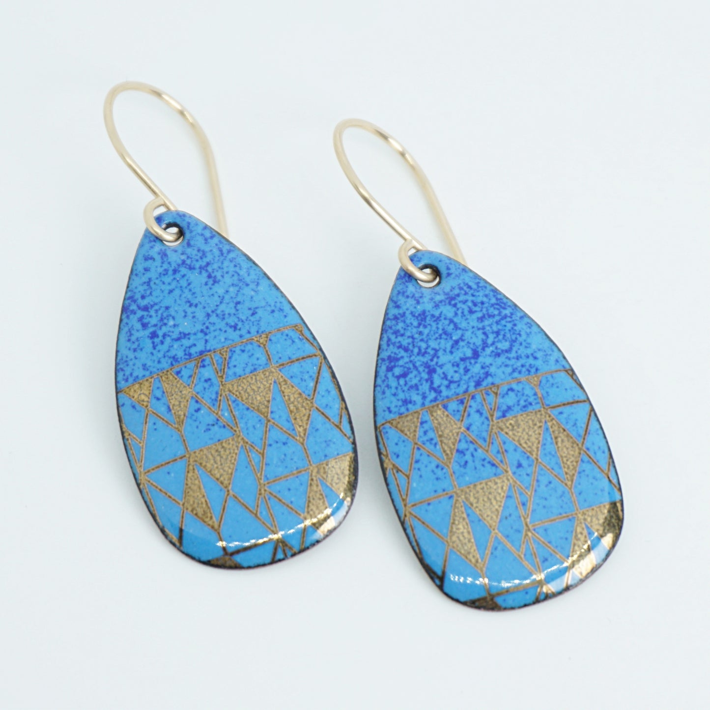 A pair of teardrop-shaped drop earrings with a blue speckled top half and a geometric gold triangle pattern on a blue lower half, hanging from thin gold hooks against a white background