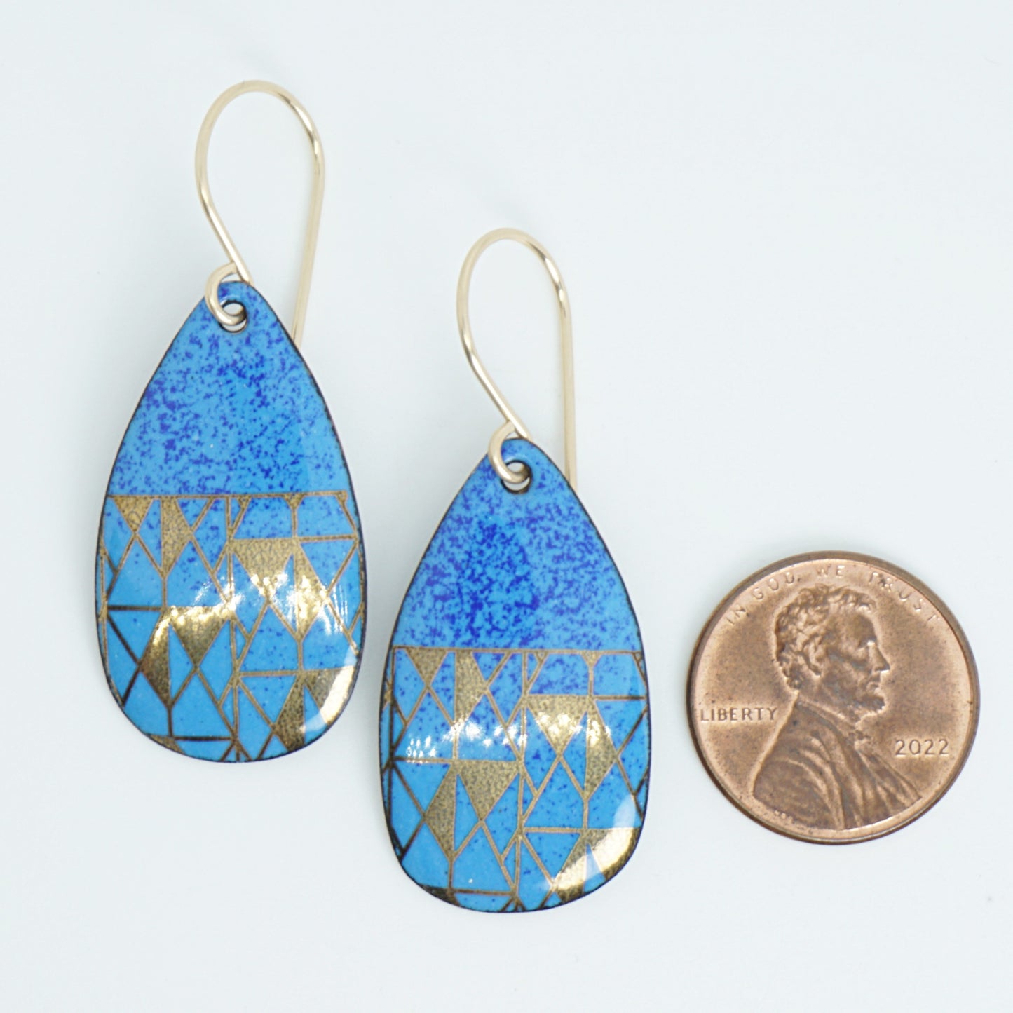 A pair of teardrop-shaped drop earrings with a textured blue enamel background and a gold geometric triangle pattern, shown next to a US penny for scale on a white background.