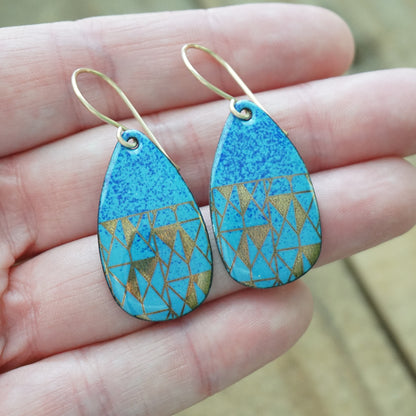 A pair of teardrop-shaped drop earrings with a textured blue top half and a geometric gold patterned bottom half, hanging from gold fishhook wires and displayed on a person's hand.