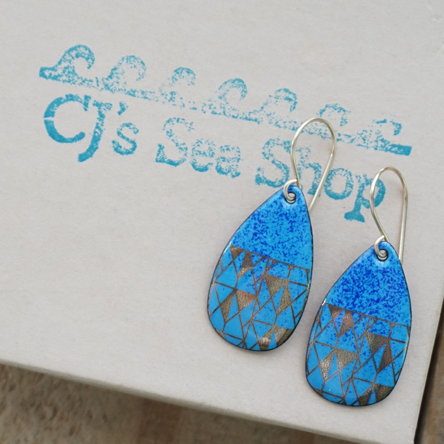 A pair of teardrop-shaped drop earrings with a blue speckled top half and a geometric gold triangle pattern on a blue lower half, hanging from thin gold hooks against a gift box