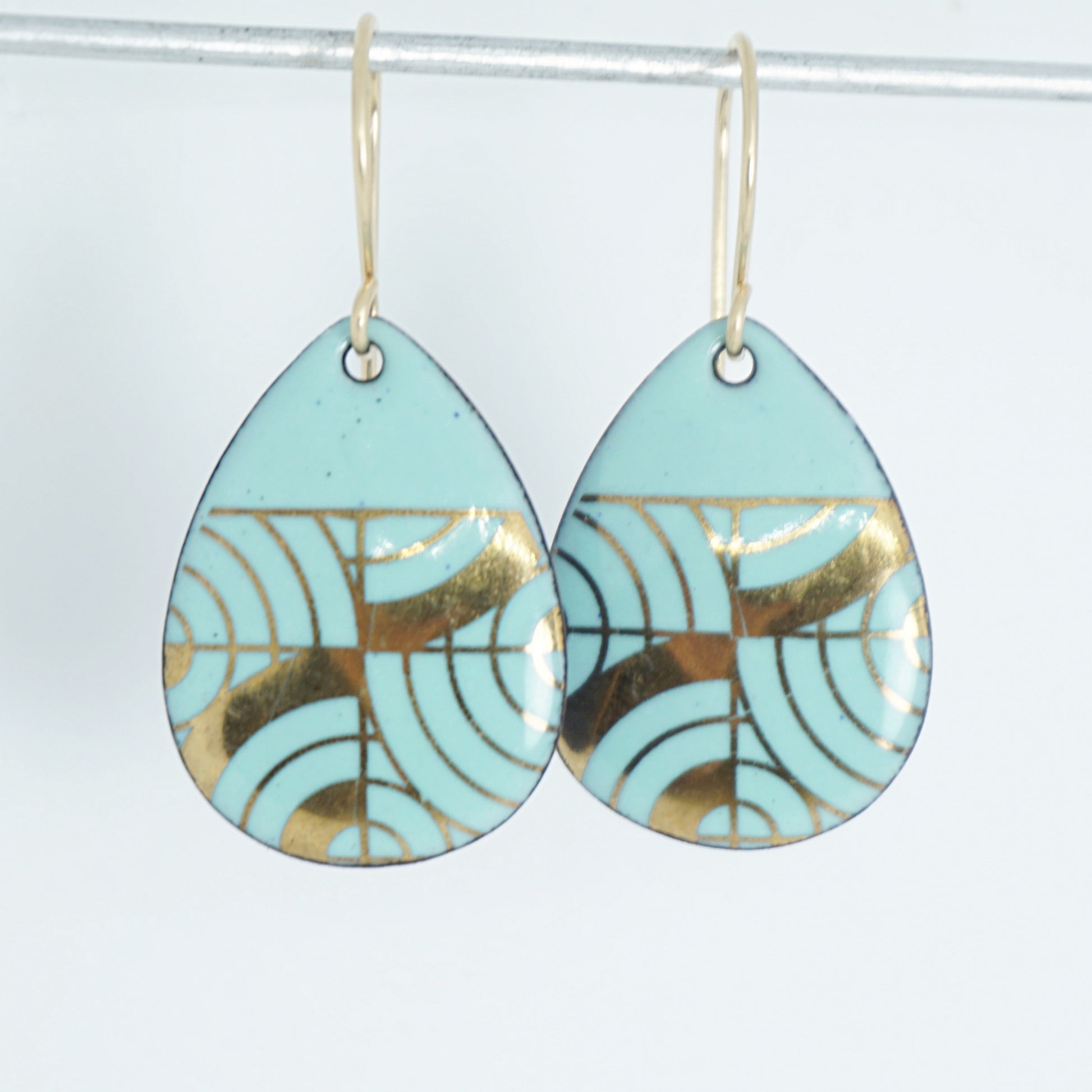 A pair of teardrop-shaped dangle earrings with seafoam green enamel and gold geometric pattern, against a white background