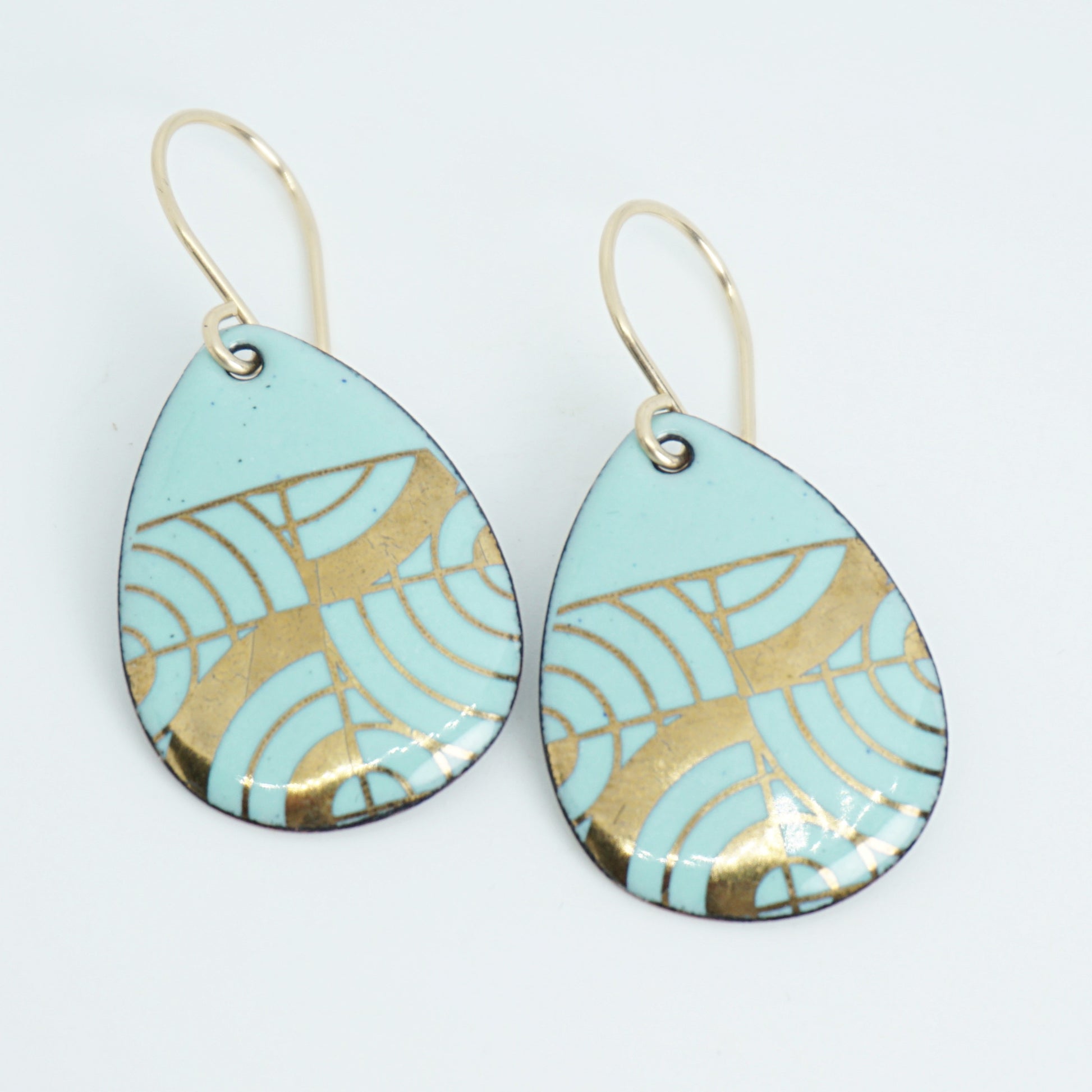 A pair of teardrop-shaped dangle earrings with seafoam green enamel and gold geometric pattern, against a white background