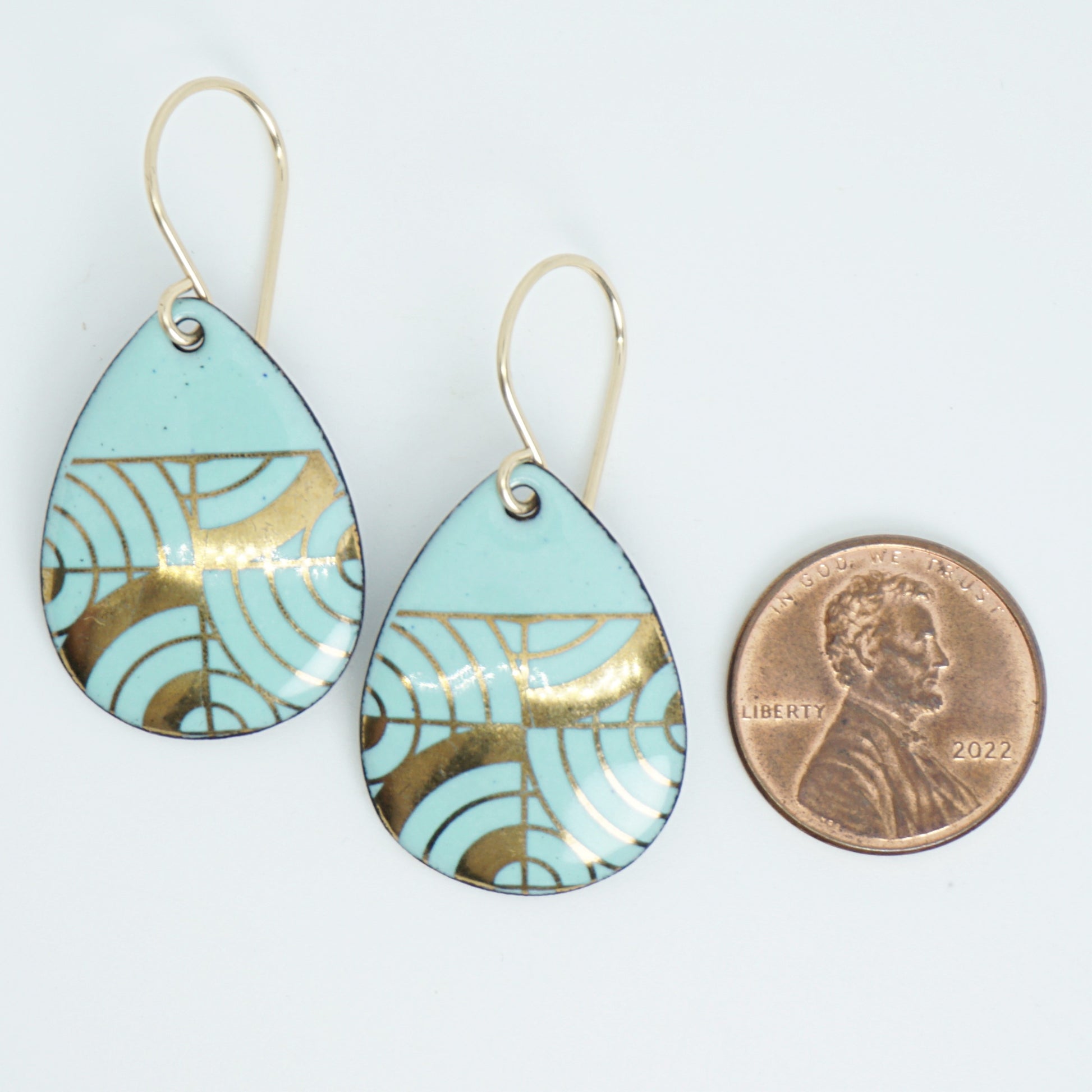 A pair of teardrop-shaped dangle earrings with seafoam green enamel and gold geometric pattern, next to a penny for scale, against a white background